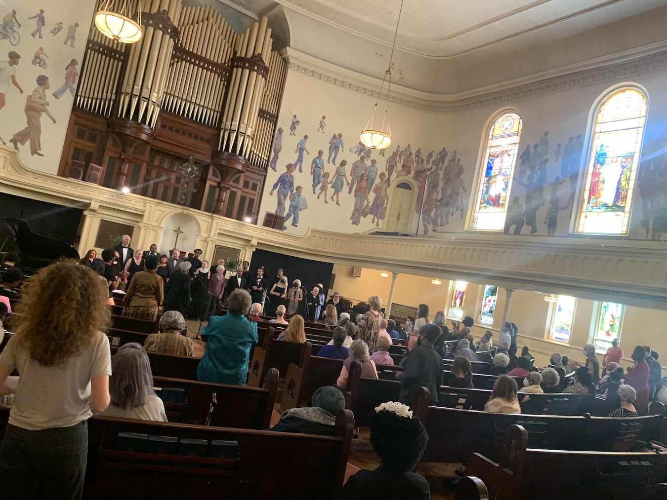 Interior of church sanctuary, crowd standing as Choir sings, light streaming in through stained glass windows.