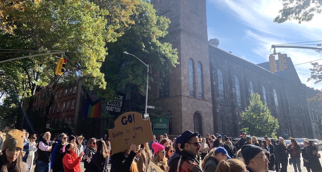 Crowd of people outside on the street with waves of marathon runners going past. Church building in the background.