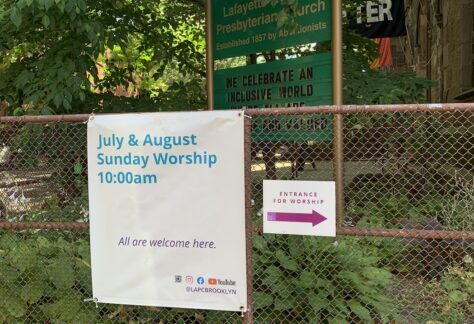 Sign on church fence with text announcing worship service starts at 10am in July and August