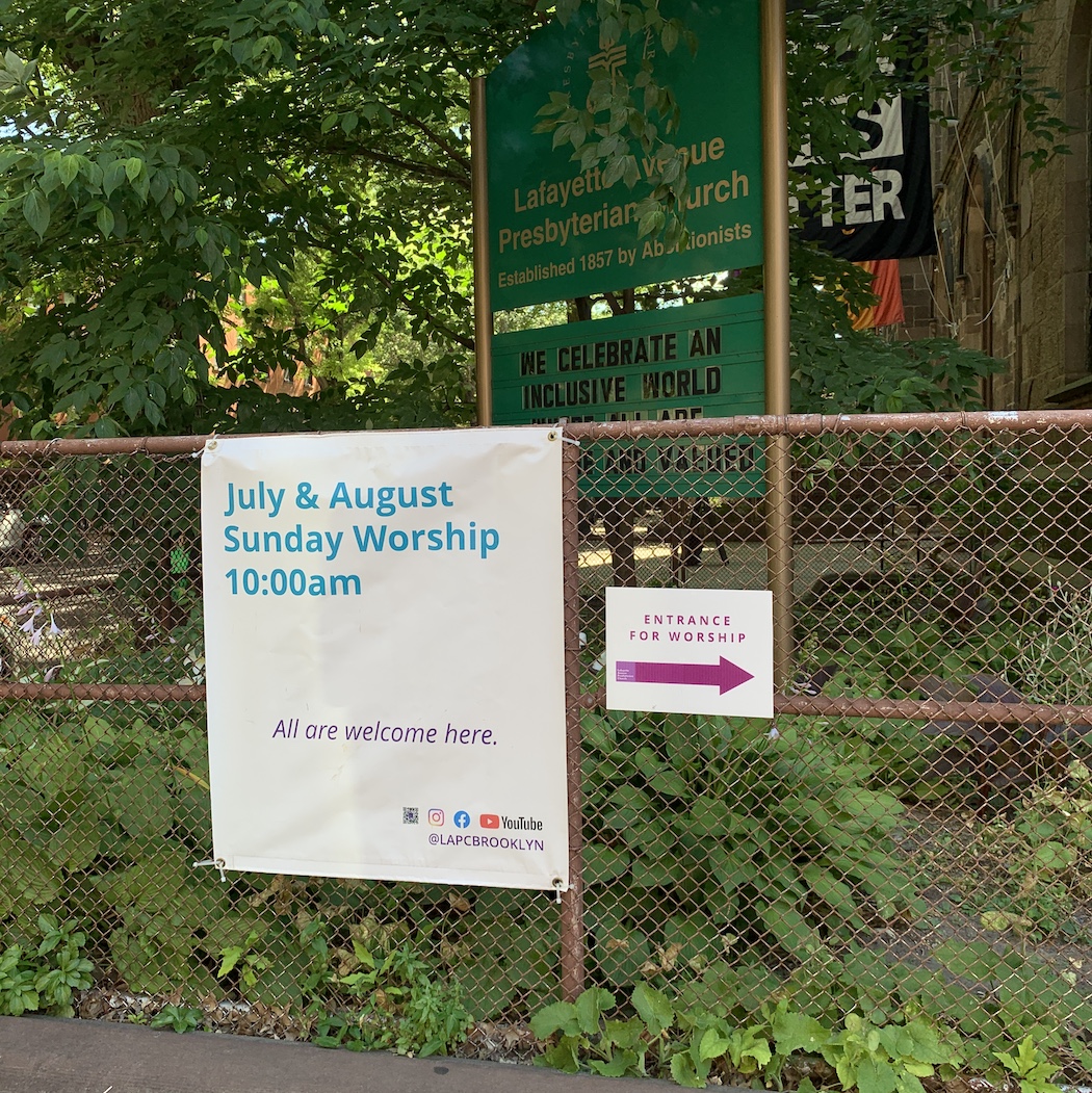 Sign on church fence with text announcing worship service starts at 10am in July and August