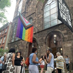 Crowd of people gathered in front of the church, with two flags flying above, one is the Pride flag, and the other for Black Lives Matter