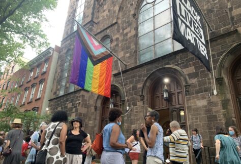 Crowd of people gathered in front of the church, with two flags flying above, one is the Pride flag, and the other for Black Lives Matter