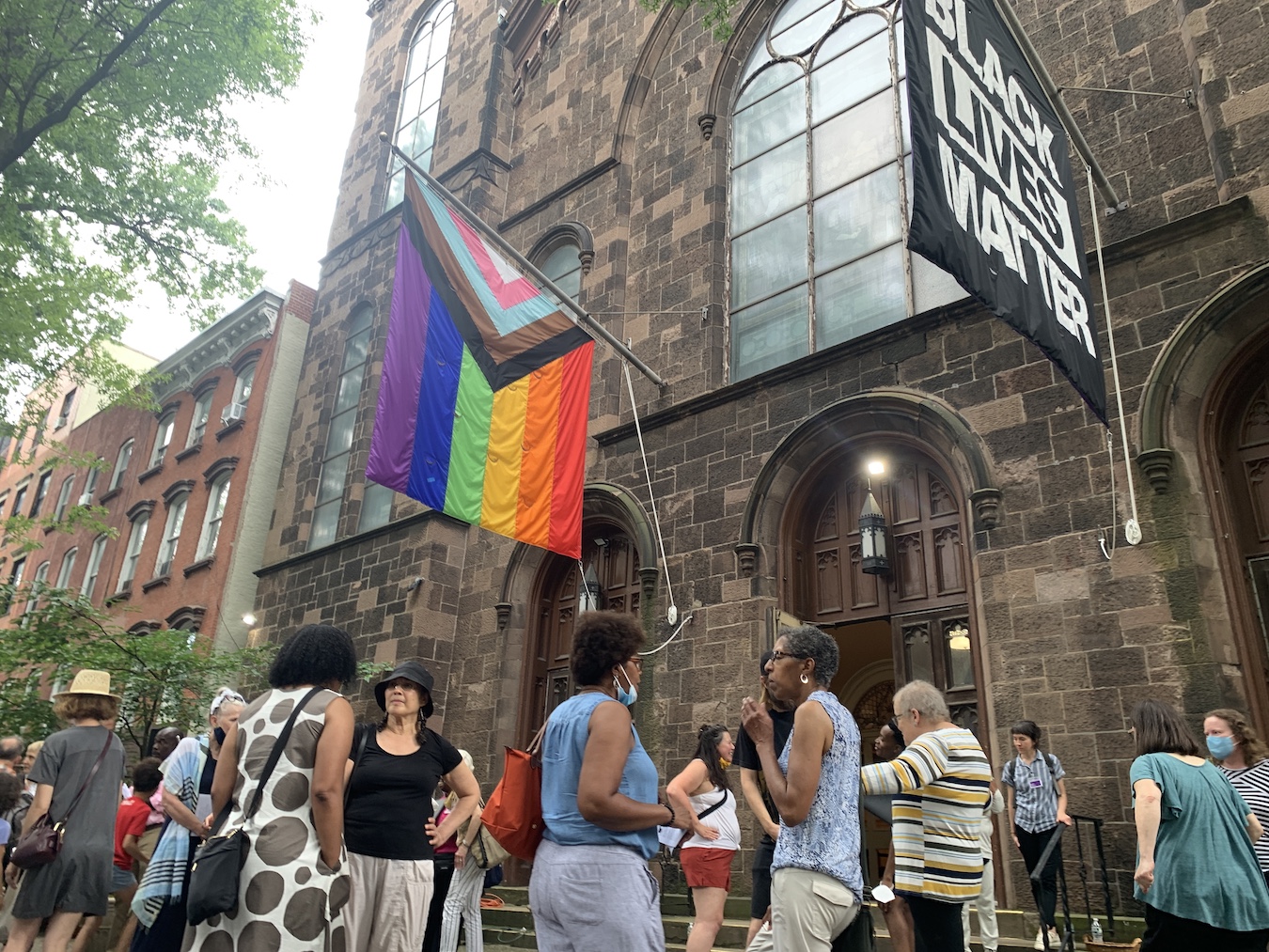 Crowd of people gathered in front of the church, with two flags flying above, one is the Pride flag, and the other for Black Lives Matter