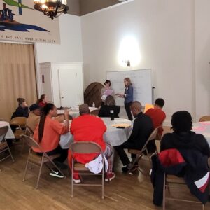 Groups of people sitting at round tables in the church's community room. Two presenters at the front.
