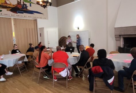 Groups of people sitting at round tables in the church's community room. Two presenters at the front.