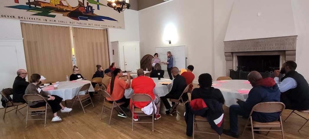 Groups of people sitting at round tables in the church's community room. Two presenters at the front.