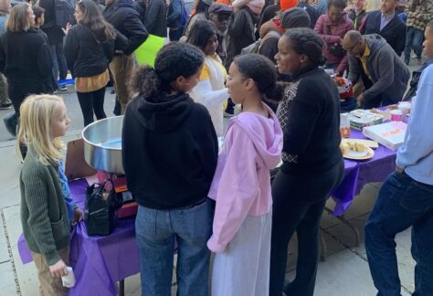 Large crowd of people in the street running the marathon, foreground a group of teens at a table of baked goods